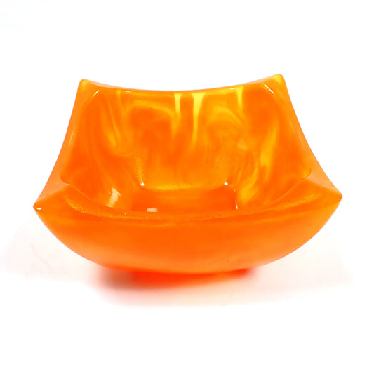 Angled view of the handmade decorative bowl. The resin is marbled bright pearly orange and semi translucent neon orange in color. The bowl is square shaped with angled pinched edges for a retro style appearance.