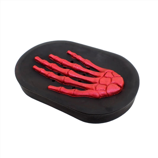 Angled view of the handmade Goth Halloween soap dish. It is oval shaped with a skeleton hand in the middle. The hand is pearly bright red and the rest of the soap dish is black. There are drain holes between the fingers.