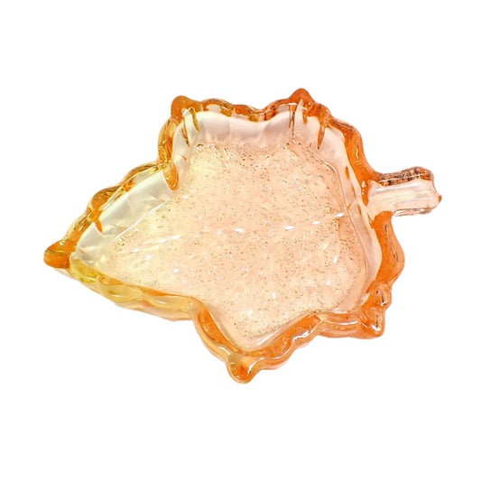 Angled view of the handmade resin leaf trinket dish. The resin is translucent orange and yellow  in color. The inside bottom of the leaf has a faceted crystal like design that sparkles in the light.