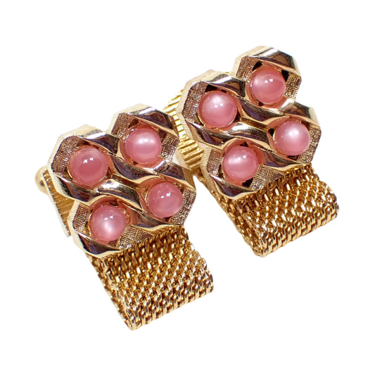 Angled view of the 1960's Mid Century vintage wrap around cufflinks. The metal is gold tone in color. There are four domed light pink moonglow lucite cabs on the front and mesh metal chain at the bottom that wraps around to the back.