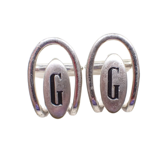 Front view of the Swank 1960's Mid Century vintage letter cufflinks. The metal is silver tone in color. They have an open arch style design over an oval that has a black painted recessed initial G in the middle.