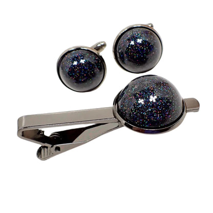 Photo of the Men's jewelry set pieces together. The metal is gunmetal gray in color. There is a tie clip and set of cufflinks. Each has a domed round resin cab with holographic black glitter that sparkles in tiny specks of rainbow colors as the light hits it.