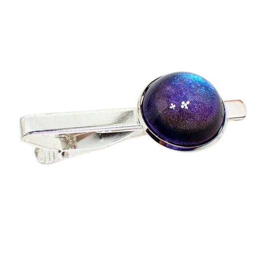 Angled view of the handmade color shift tie clip. The metal is silver tone in color. There is a domed round resin cab at the end of the bar that shifts from blue to purple with other hints of colors as the light hits it.