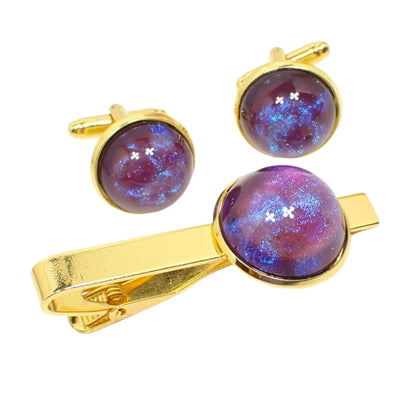 Angled view of the handmade color shift men's jewelry set. The metal is gold tone in color. There is a tie clip and pair of cufflinks in the photo. Each has a domed round resin cab that has iridescent pink and swirls of blue that shifts to purple as the light hits them.