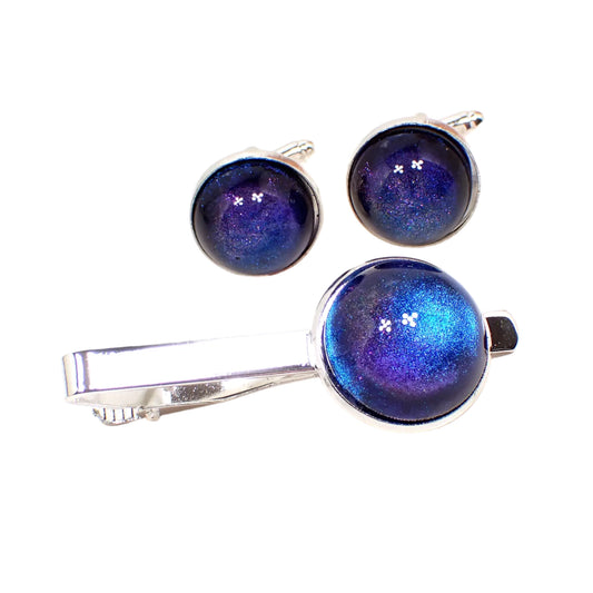 Angled view of the color shift men's jewelry set. There is a tie clip and set of cufflinks with a silver tone color metal setting. They each have domed round resin cabs with color shift design that goes from blue to purple depending on how the light hits them.