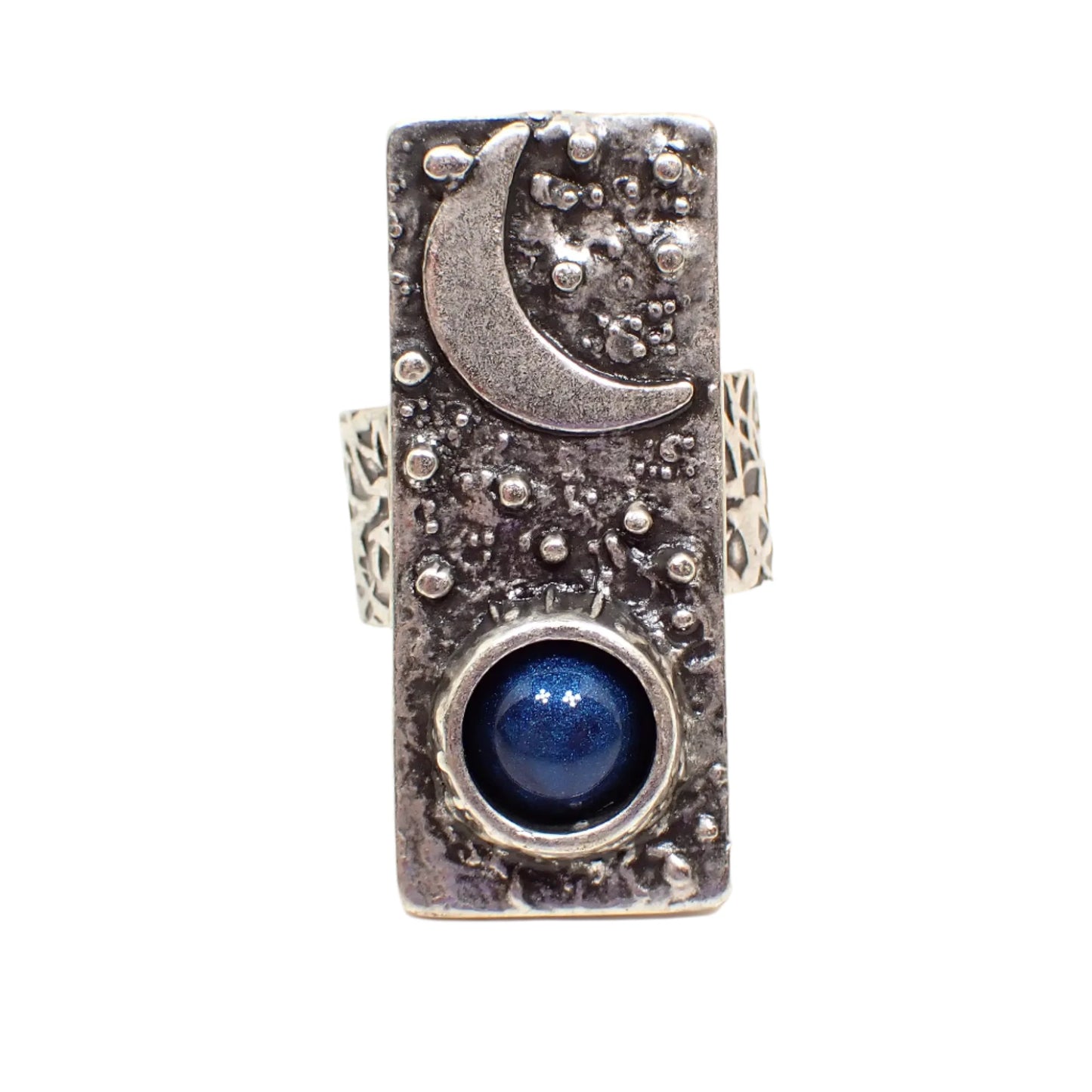 Front view of the handmade adjustable ring. The metal is antiqued silver tone in color. The top of the ring has a long rectangle bar shape that has a raised moon at the top and raised dots for stars. The bottom has a domed round resin cab in dark denim blue color.