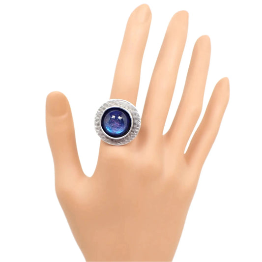 Photo of the handmade adjustable ring shown on a mannequin hand. It is round and silver tone in color. The edge has a hammered design. The middle has a domed round resin cab that color shifts from blue to purple as the light hits it.