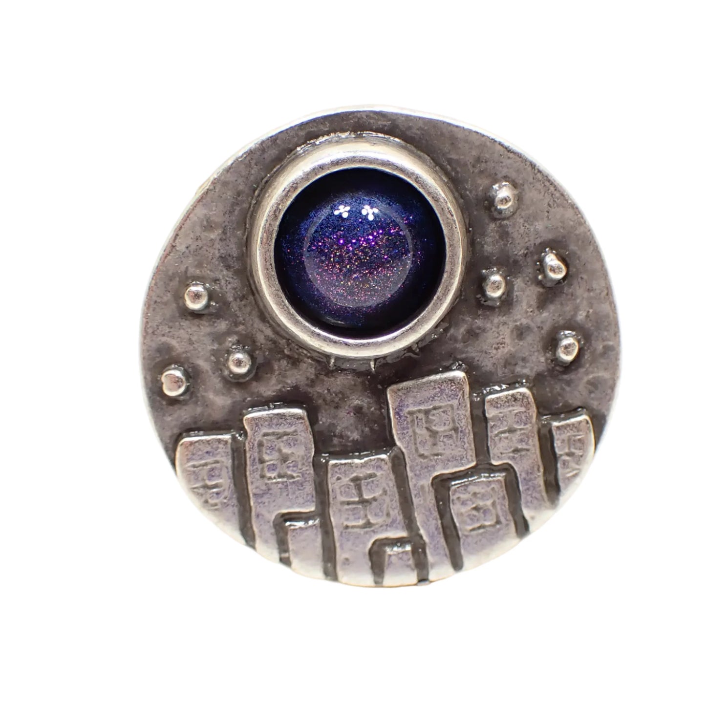 Front view of the handmade adjustable ring. The metal is antiqued silver tone in color. The top of the ring is round and has a raised design of city buildings with dotted stars overhead. There is a domed round resin cab at the top that is color shift from blue to purple depending on how the light hits it.