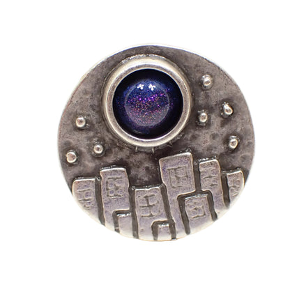 Front view of the handmade adjustable ring. The metal is antiqued silver tone in color. The top of the ring is round and has a raised design of city buildings with dotted stars overhead. There is a domed round resin cab at the top that is color shift from blue to purple depending on how the light hits it.