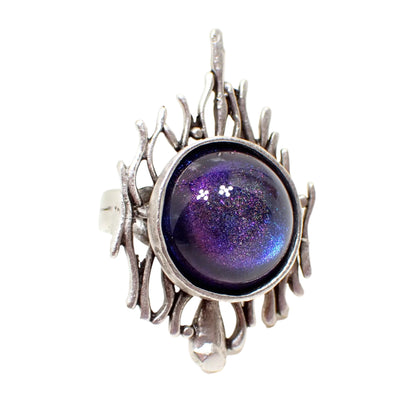 Angled front view of the Brutalist style handmade adjustable ring. The metal is silver tone in color. It has a Open style design with a larger sized domed round resin cab in the middle. The color shift resin cab goes from blue to purple as the light hits it.