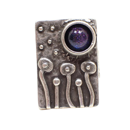 Front view of the handmade adjustable ring. It is rectangle shaped on top and has antiqued silver tone color metal. The front has a primitive style raised design depicting flowers and mushrooms with rounded tops on curvy stems with dot stars overhead. There is a domed round resin cab at the top right that color shifts from blue to purple depending on how the light hits it.