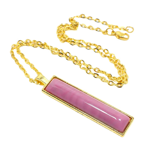 Angled view of the handmade resin bar pendant necklace. It has a long rectangle bar shape with a handmade domed resin cab that is marbled rose pink in color. The metal setting, chain, and lobster claw clasp are gold tone in color.