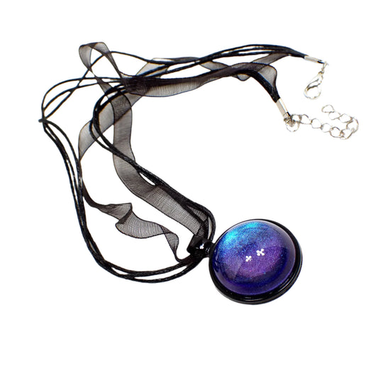 Angled view of the handmade color shift round resin pendant necklace. The metal is coated black in color. The round pendant has a domed round cab with a color shift design that goes from blue to purple depending on how the light is hitting it. The necklace part has black organza and strands of waxed cord.