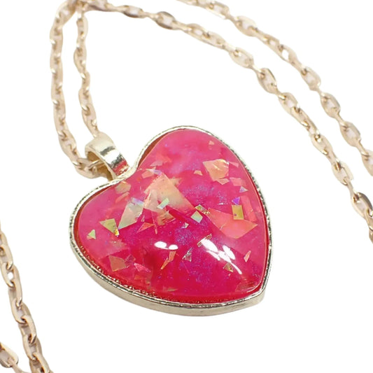 Enlarged view of the handmade resin heart pendant necklace. The metal is gold tone in color. It has an oval cable link chain and a heart shaped metal pendant. The pendant has a domed resin cab in a bright pearly pink color that has iridescent flakes embedded in it that give off flashes of opalescent like color.