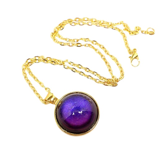 Angled view of the handmade color shift round resin pendant necklace. The metal is gold tone in color. The round pendant has a domed round cab with a color shift design that goes from blue to purple depending on how the light is hitting it.