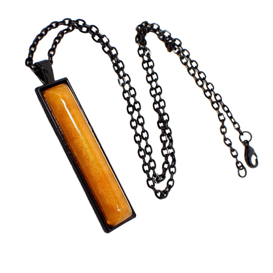 Angled view of the Halloween bar pendant necklace. The chain and pendant setting are black coated. The bar pendant is long rectangle shaped and has a domed resin cab that is pearly orange in color. There is a lobster claw clasp at the end of the cable chain.