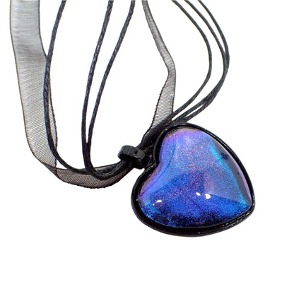 Enlarged view of the handmade color shift blue purple resin black heart pendant necklace. There is a strand of black organza and strands of waxed cord with a heart shaped pendant at the bottom. The pendant has black coated metal and a resin cab that has a color shift from blue to purple depending on how the light hits it.