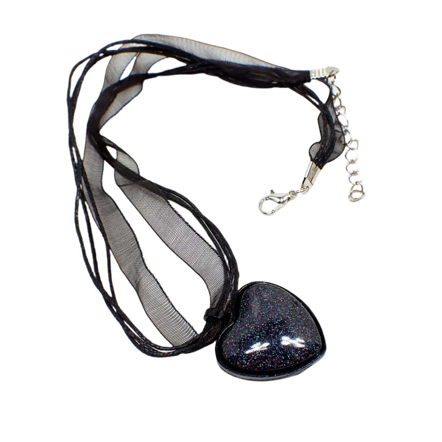 Angled view of the handmade Goth pendant necklace. The necklace part has a strand of black organza and strands of waxed cord. There is a silver tone extender chain and lobster claw clasp at the end. The pendant is heart shaped with black coated metal and a resin cab that has holographic black glitter resin.