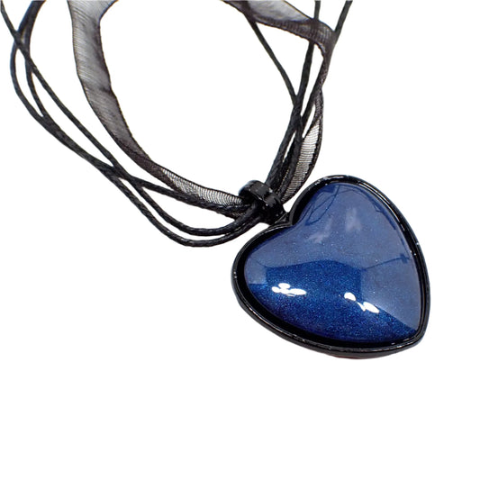 Enlarged view of the handmade resin Goth pendant necklace. The necklace has a strand of organza and strands of waxed cord. There is a heart shaped pendant with black coated metal. The resin cab is a dark denim blue in color.