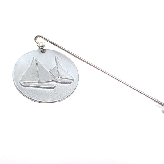 Enlarged view of the retro vintage stick pin. The metal is silver tone in color. The top has a round dangling matte pewter charm with a raised design of two sailboats on it.
