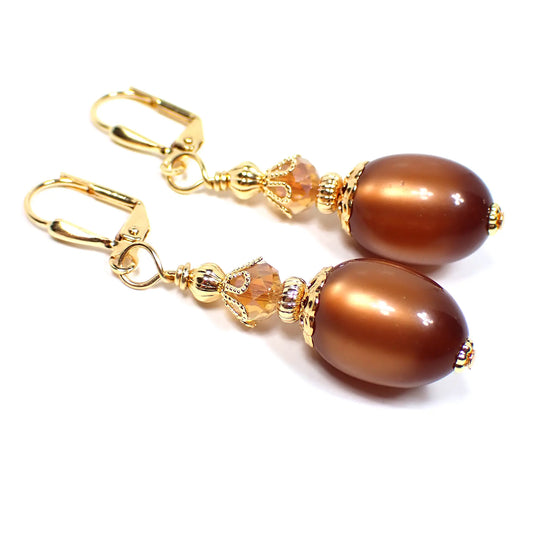 Angled view of the handmade earrings with vintage moonglow lucite beads. The metal is gold plated in color. There are faceted glass crystal beads at the top with shades of orange and peach. The bottom lucite beads are oval shaped and are a golden brown color with highlights of lighter brown and orange where the light is hitting them.