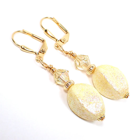 Angled view of the handmade drop earrings with vintage acrylic glitter beads. The metal is gold plated in color. There are light yellow faceted crystal glass beads at the top. The bottom acrylic beads are a twisted oval like shape and light yellow in color with tiny flecks of iridescent glitter.
