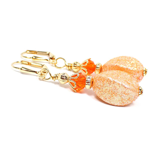 Angled view of the handmade drop earrings with vintage acrylic glitter beads. The metal is gold plated in color. There are bright orange faceted crystal glass beads at the top. The bottom acrylic beads are a twisted oval like shape and lighter shade of orange in color with tiny flecks of iridescent glitter.