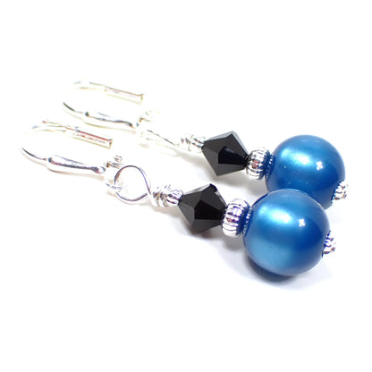 Angled view of the handmade small drop earrings with vintage moonglow lucite beads. The metal is bright silver in color. There is a black faceted glass crystal bead at the top. The bottom moonglow lucite bead is round ball shaped and teal blue in color.