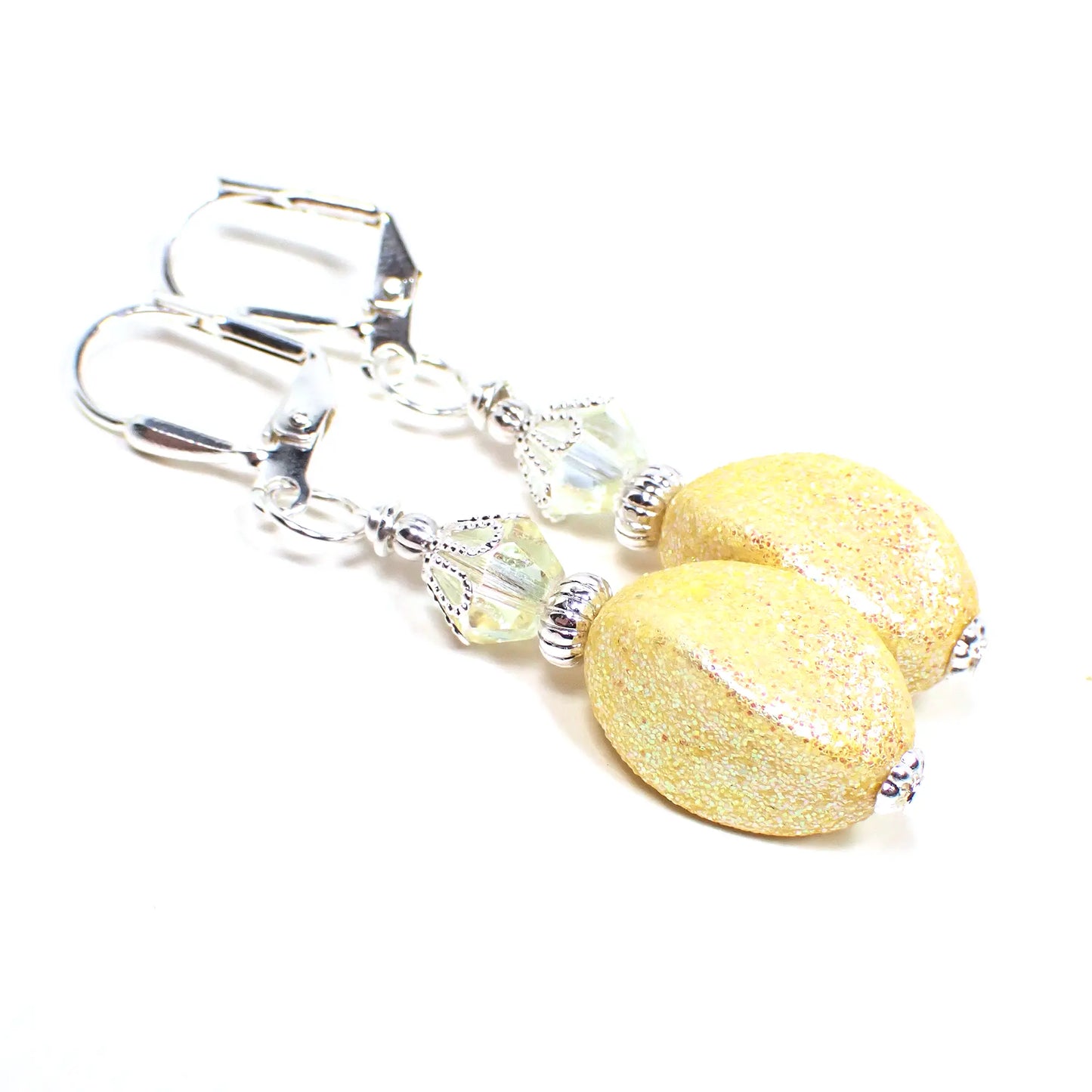 Angled view of the handmade drop earrings with vintage acrylic glitter beads. The metal is bright silver in color. There are light yellow faceted crystal glass beads at the top. The bottom acrylic beads are a twisted oval like shape and light yellow in color with tiny flecks of iridescent glitter.
