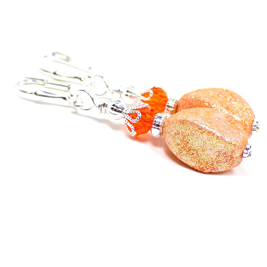 Angled view of the handmade drop earrings with vintage acrylic glitter beads. The metal is bright silver in color. There are bright orange faceted crystal glass beads at the top. The bottom acrylic beads are a twisted oval like shape and lighter shade of orange in color with tiny flecks of iridescent glitter.