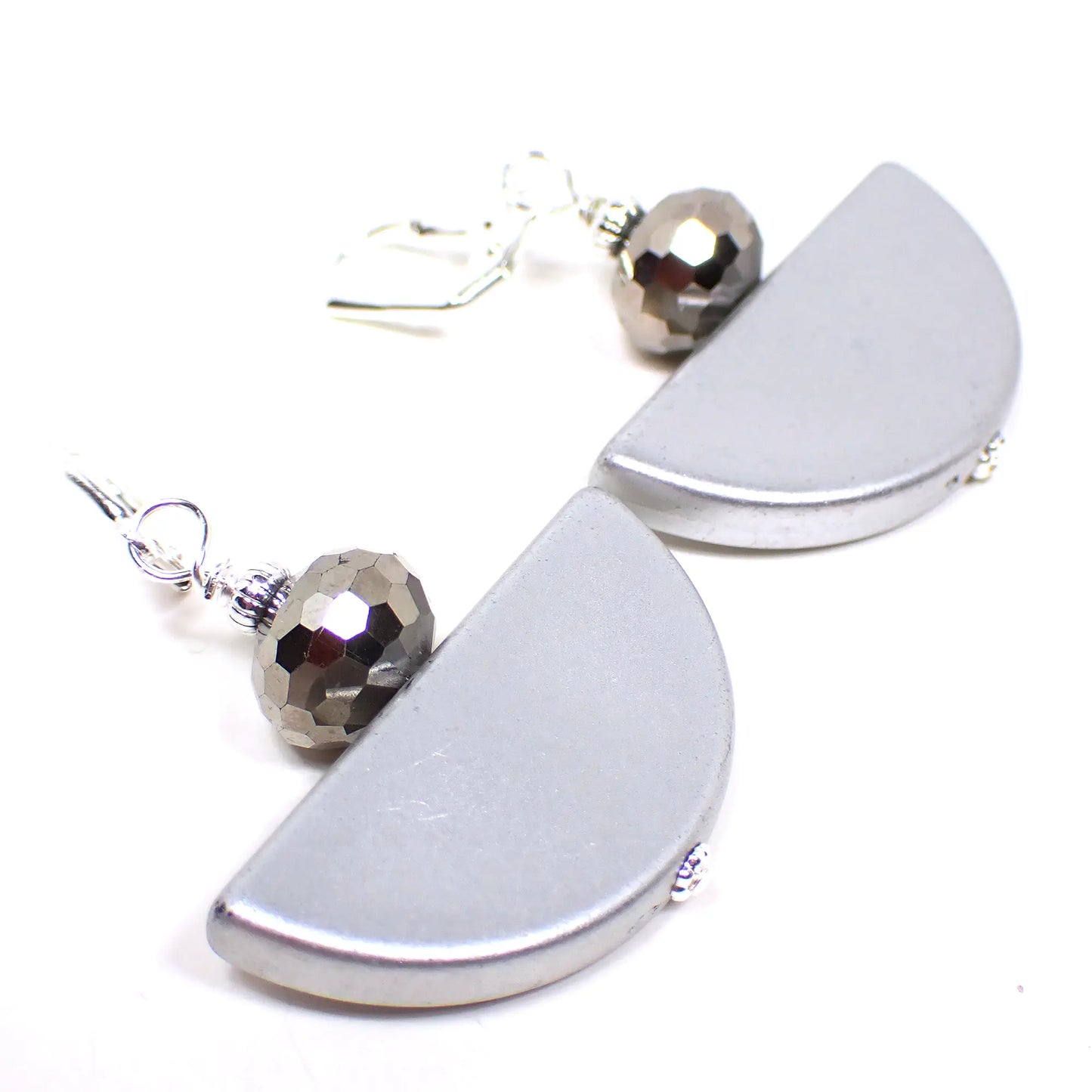 Angled view of the handmade half moon earrings. The metal is bright silver in color. There are faceted round metallic silver colored glass beads on top. The bottom beads are vintage acrylic beads that are half moon shaped and matte metallic silver in color.
