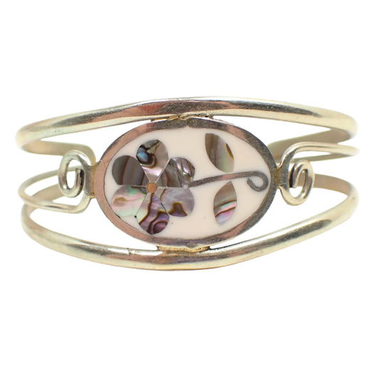 Front view of the retro vintage Alpaca Mexico cuff bracelet. The metal is silver tone in color. There is an oval on the front that has white enamel and inlaid pieces of iridescent abalone shell in the shape of a flower.