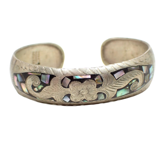 Front view of the vintage Alpaca cuff bracelet. The metal is antiqued silver tone in color. It has an etched flower and leaves design on the front surrounded by black enamel that has small inlaid pieces of iridescent abalone shell.