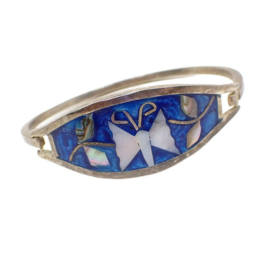 Angled front view of the Alpaca Mexico marked vintage hinged bangle bracelet. The metal is antiqued silver tone in color. The front is wider in the center with pearly ocean blue enamel, a mother of pearl shell butterfly in the middle, and abalone shell leaves on either side.