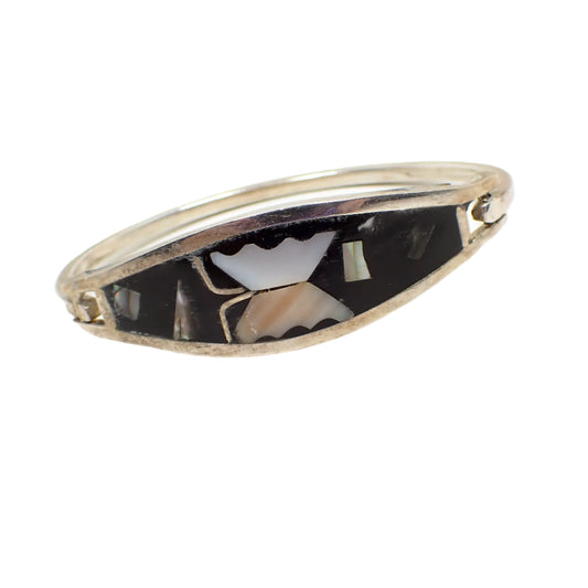 Angled front view of the Alpaca vintage hinged bangle bracelet. The metal is silver tone in color. The front is black enameled. It has a mother of pearl shell butterfly as well as a few other shell pieces throughout. 