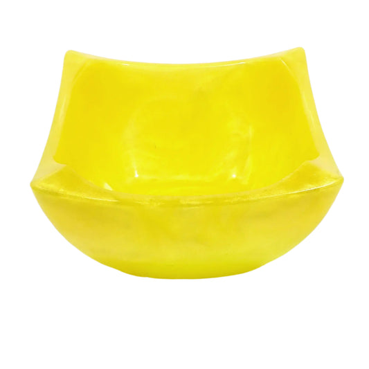 Angled side view of the handmade resin square decorative bowl. The resin is shiny and opaque in a pearly bright yellow color. The bowl has an atomic style square design with flared out pinched edges and a tapered bottom. 