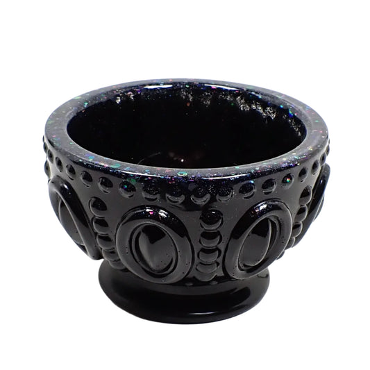 Angled side view of the handmade footed bowl. It's round with an oval and dot pattern on the outside. The resin is shiny black and there is iridescent glitter at the top.