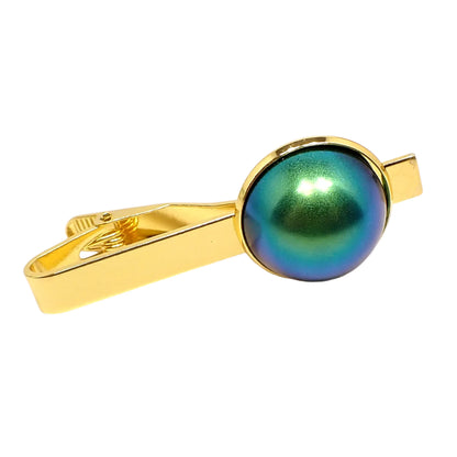 Angled front view of the tie clip. The metal is gold tone in color. There is a  large domed round resin cab at the end with matte metallic shades of blue and green.