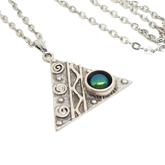 Enlarged close up of the pendant. It is antiqued silver tone and shaped like a triangle. There are swirls, dots, a curvy line between straight lines, and a round domed resin cab. The cab has shades of blue and green depending on how the light is hitting it.