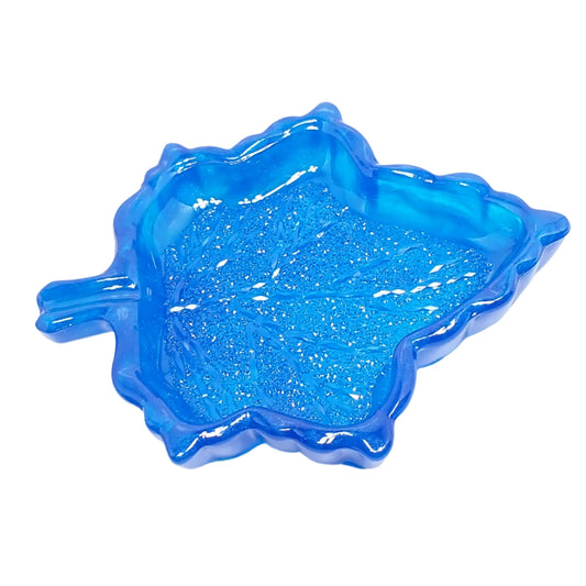 Angled view of the blue leaf trinket dish. It has neon blue with pearly blue around the edges for an overall bright blue appearance. It's shaped like a maple leaf and has small facets on the inside bottom area giving it a crystal like look that sparkles when the light hits it.