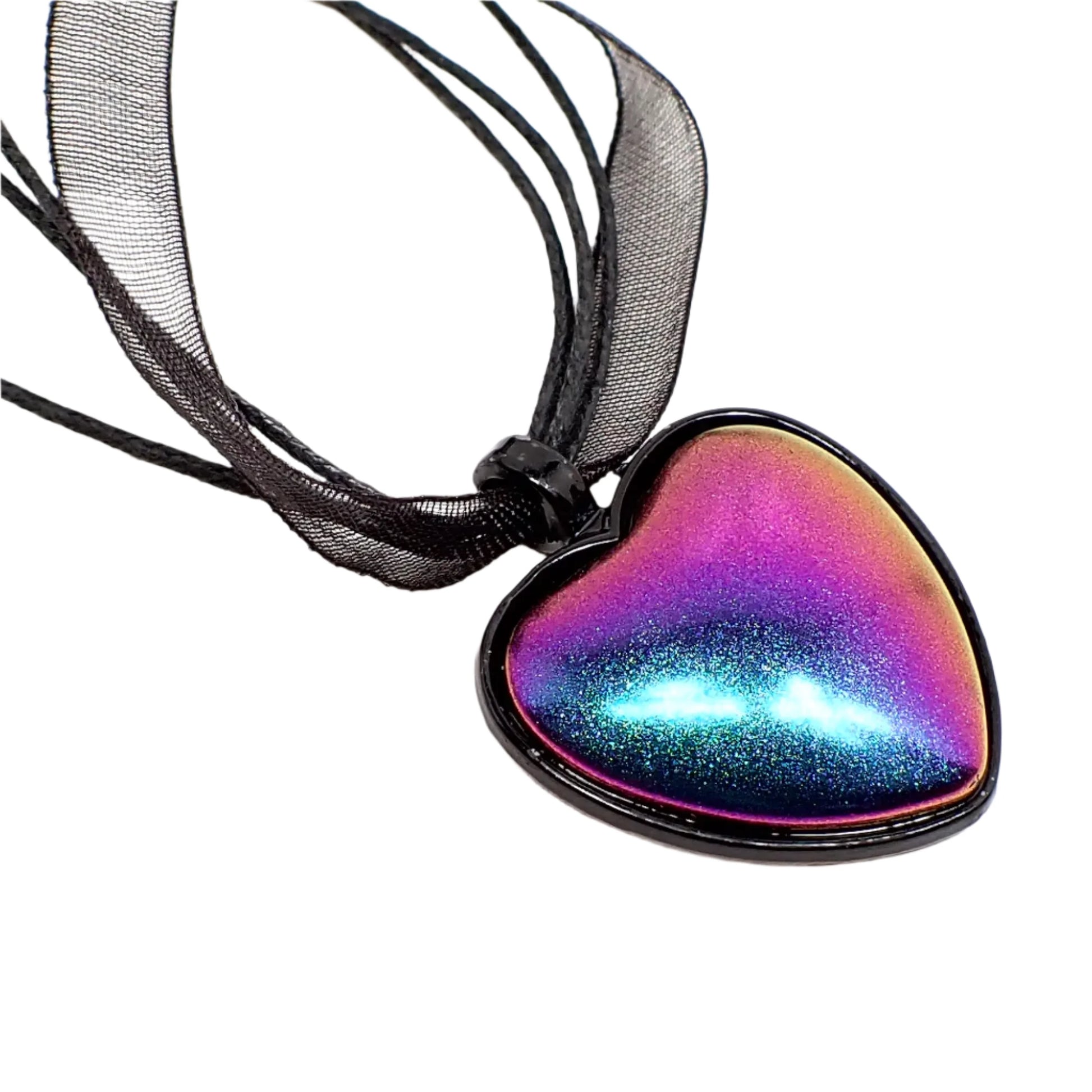 Enlarged close up of the pendant. The setting is a black enameled heart shape and has a color shift cab. Shades of bright matte metallic blue, pink, and purple can be seen. There is a strand of organza and strands of waxed cord going through the top loop of the pendant.