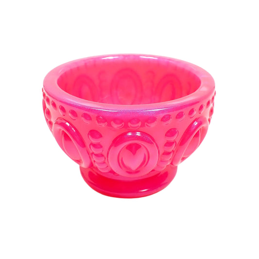 Angled side view of the handmade bright pink resin footed decorative bowl. It is round with large raised ovals around the outside that have rows of raised dots above, below, and in between the ovals. The footed bottom is flared, round, and smooth. The resin is pearly and opaque in a bright hot pink color. 