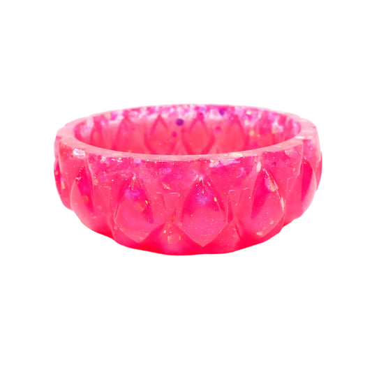 Angled side view of the handmade resin ring dish. It is short and round with a carved like diamond shape design all the way around the outside. The resin is shiny in a pearly bright pink color with iridescent chunky pink glitter around the top. 