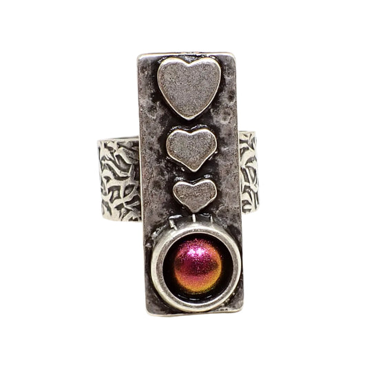 Front view of the adjustable heart ring. The metal is antiqued silver in color. The top of the ring is rectangle bar shaped. There are three hearts going down in size on the top part of the ring. The bottom has a domed round color shift resin cab that shows mostly pink but has flashes of orange and yellow seen around the edge in this photo.