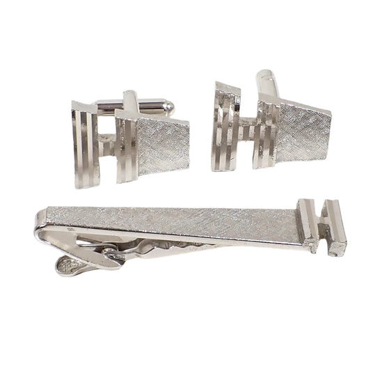 Angled view of the entire Dante Mid Century vintage mens jewelry set. The metal is silver tone in color with a matte brushed texture design. There is a pair of cufflinks and tie clip in the image. Each piece has a angled and flared atomic style design with an initial letter H at the end. The initial has a diamond cut etched design and is shiny.