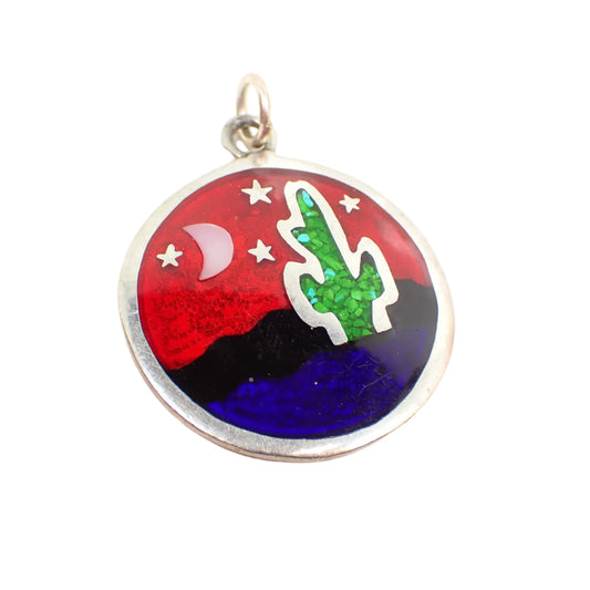 Front view of the vintage Desert Night pendant. It is round with a desert, cactus, moon, and stars design. The moon has inlaid mother of pearl and the cactus has inlaid green stone chips. There is red, black, and blue enamel on the rest of the piece. The top has a loop and jump ring, but there is no chain. Metal is silver tone in color.