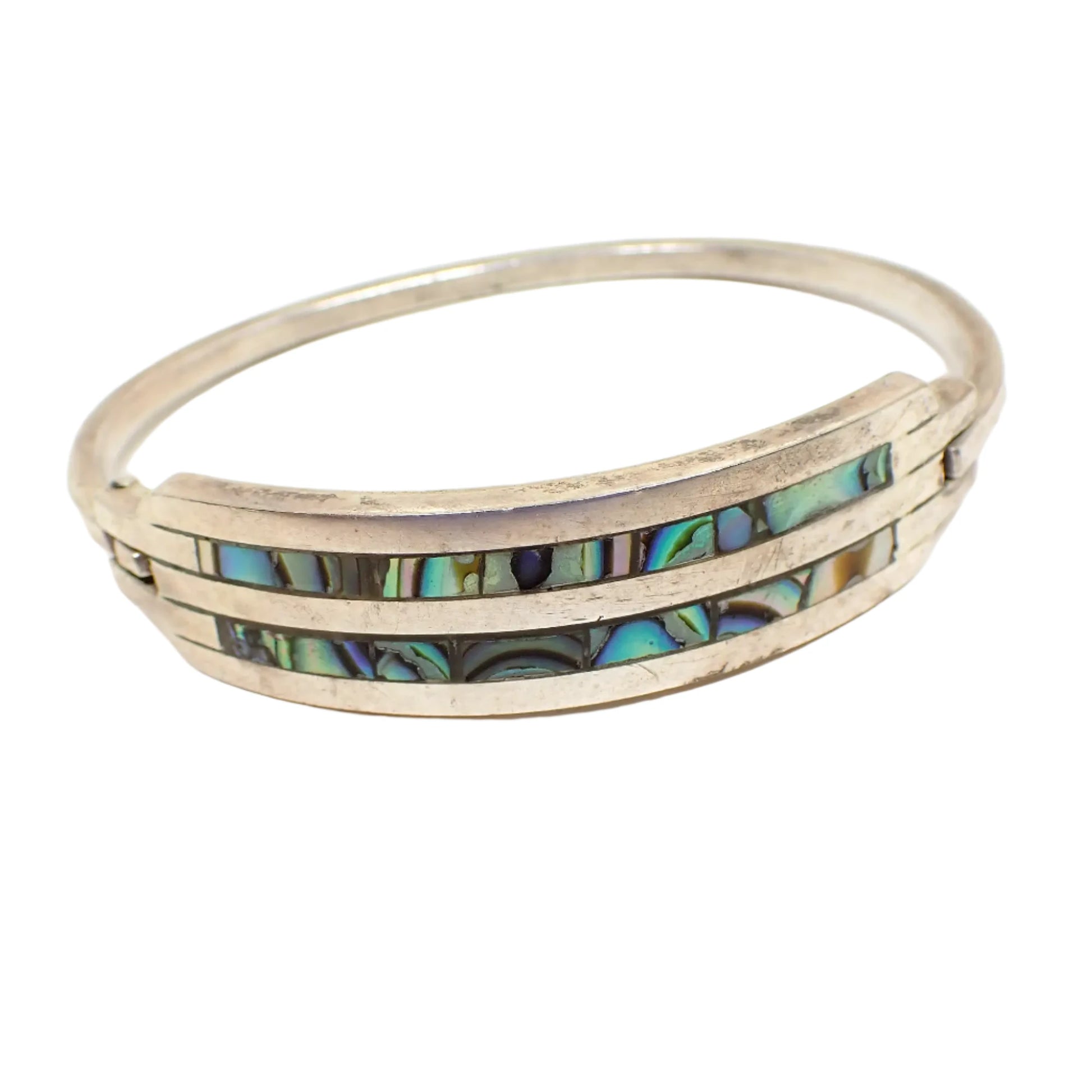 Angled view of the vintage Alpaca double row abalone bangle bracelet. The metal is silver tone in color. There are two rows curved around the front of inlaid abalone shell that is iridescent multi color. There is a hinge on one side of the bracelet and a snap clasp on the other.