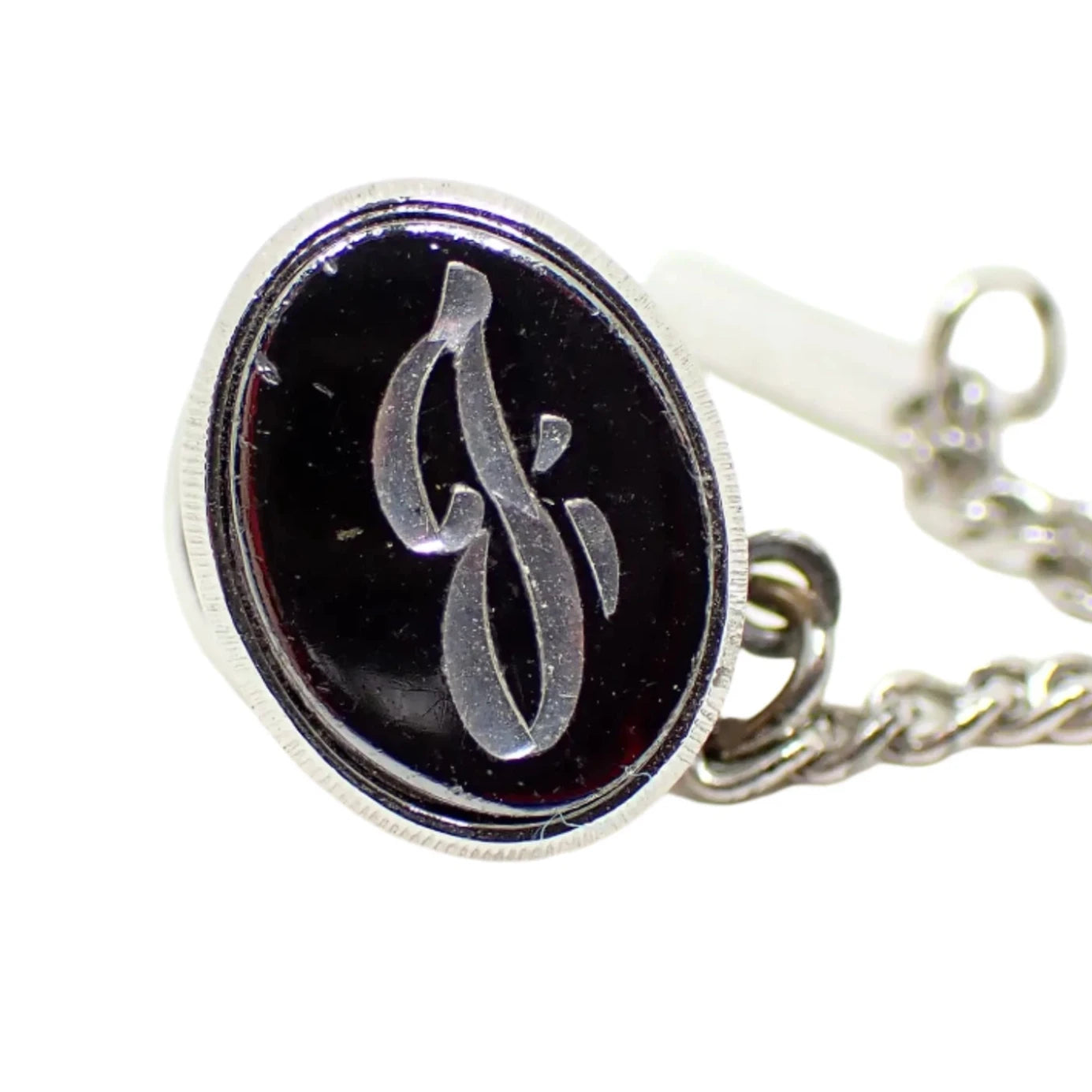 Enlarged view of the Mid Century vintage initial tie tack. It is oval with metallic gray faux hematite on the front and silver tone color metal on the rest of the piece. There is a letter J engraved in the center.