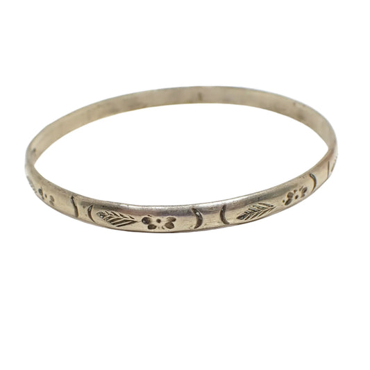 Angled view of the flower and leaf bangle bracelet. It is antiqued silver in color. The bangle is thinner style with rounded edges and a stamped leaf and flower design all the way around it. 