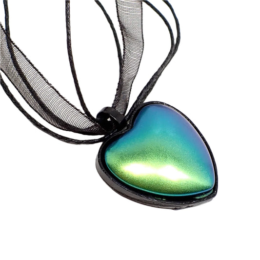 Enlarged close up of the pendant. The heart setting is black and the cab has shades of green and blue depending on how the light is hitting it. Organza and strands of cord can be seen going through the pendant loop.